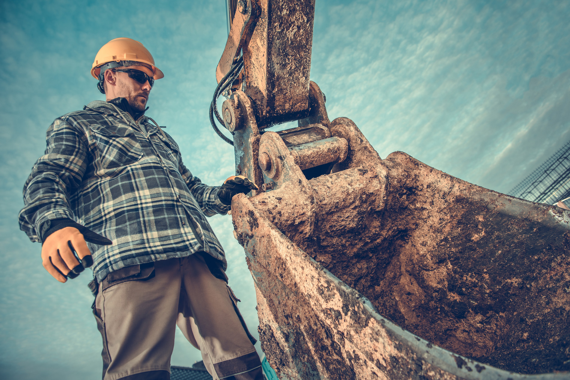 Construction,Site,And,The,Heavy,Duty,Machinery.,Ground,Works,Excavation. A construction worker with a large excavator bucket. He is working hard. Upward angle photo.